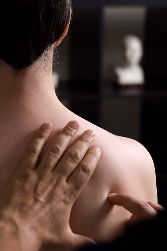 A person's shoulder being massaged in a dimly lit room. The background is slightly blurred, with a white sculpture visible on a shelf. Calm atmosphere.