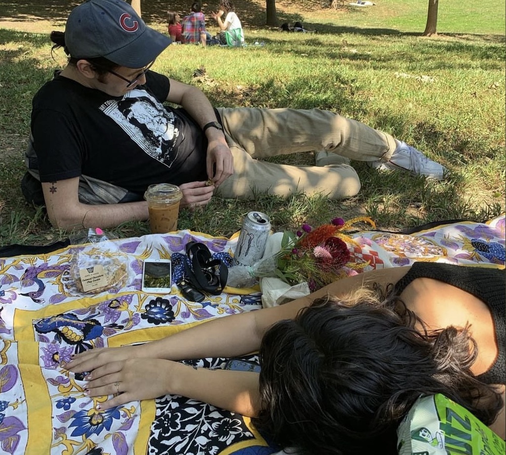 bml wears a cubs hat and lays in the grass with a coffee by their side, looking down. cb sprawls out across a blanket with their back to the camera