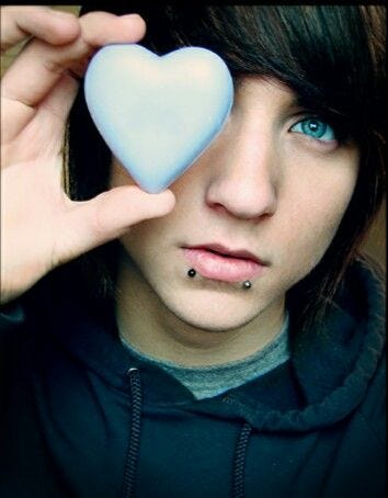 alex evans, scene boy icon with snakebite piercings and swoopy hair, holds a blue heart up over one of his piercing blue eyes.