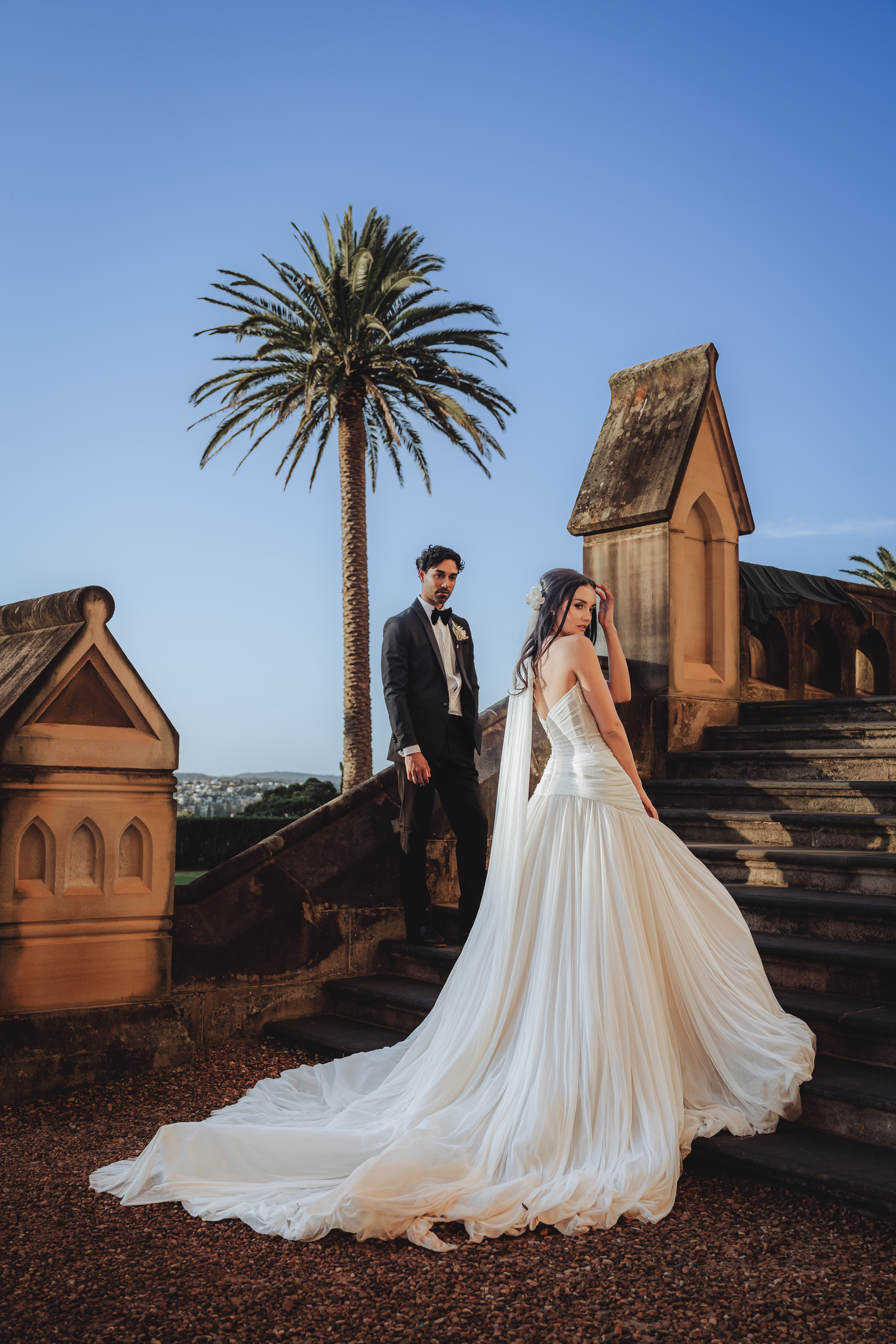 Bride in a flowing white gown and groom in a black tuxedo standing on stone steps with a palm tree and blue sky in the background.