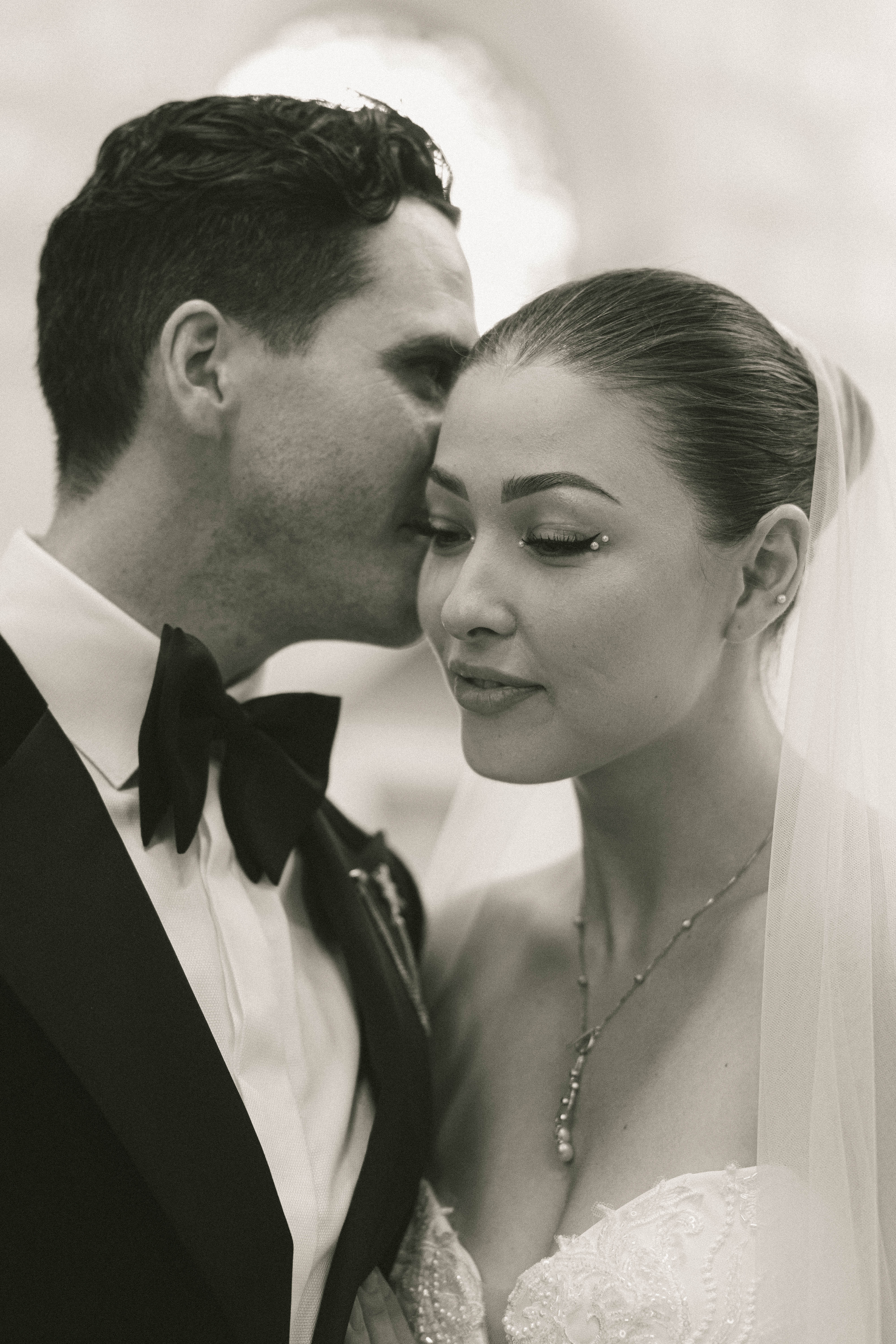 Groom in tuxedo gently whispering to bride wearing a veil and pearl necklace in a tender moment.