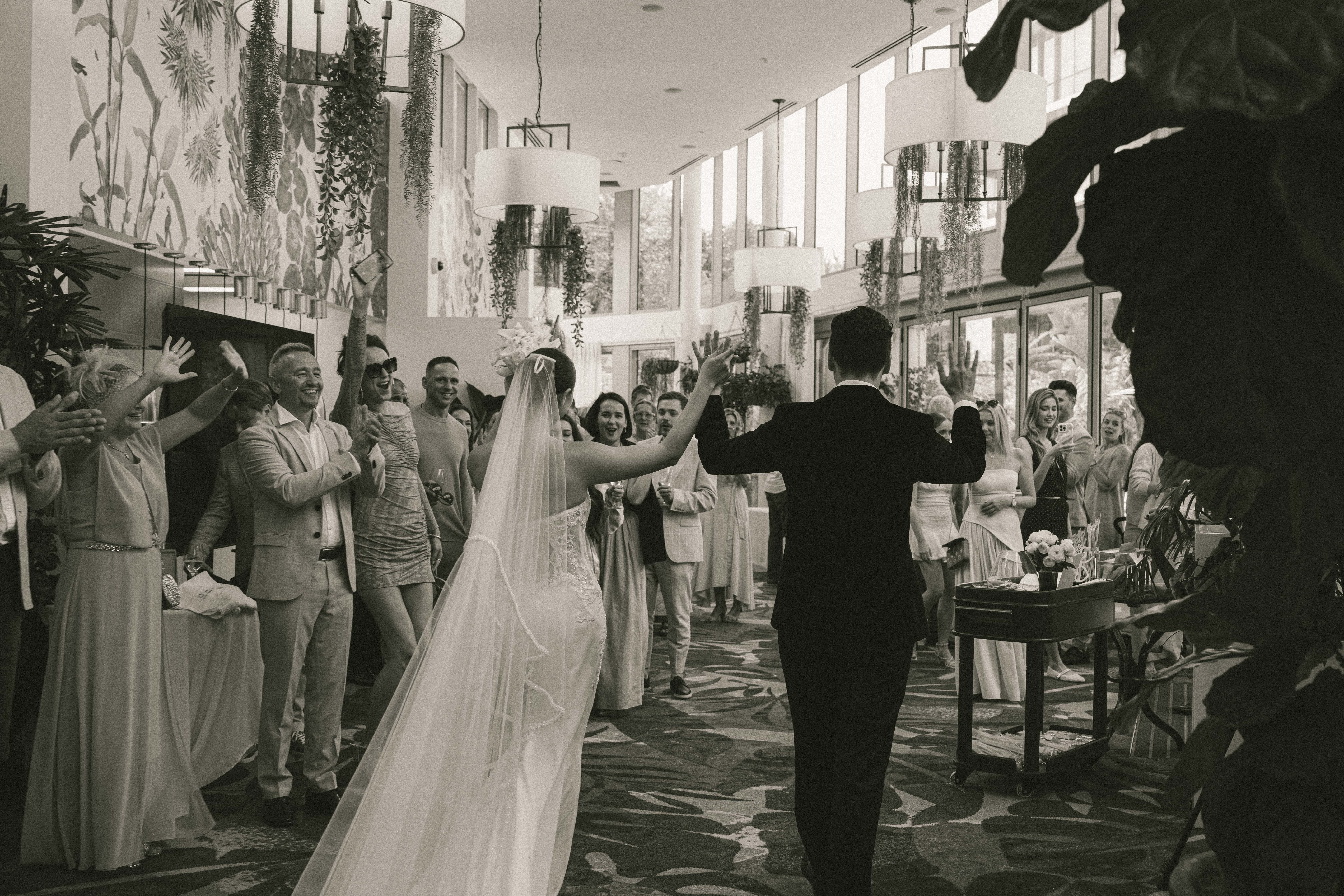 Black and white photo of a bride and groom walking hand in hand down an aisle lined with smiling guests with raised hands inside a bright, plant-decorated venue.