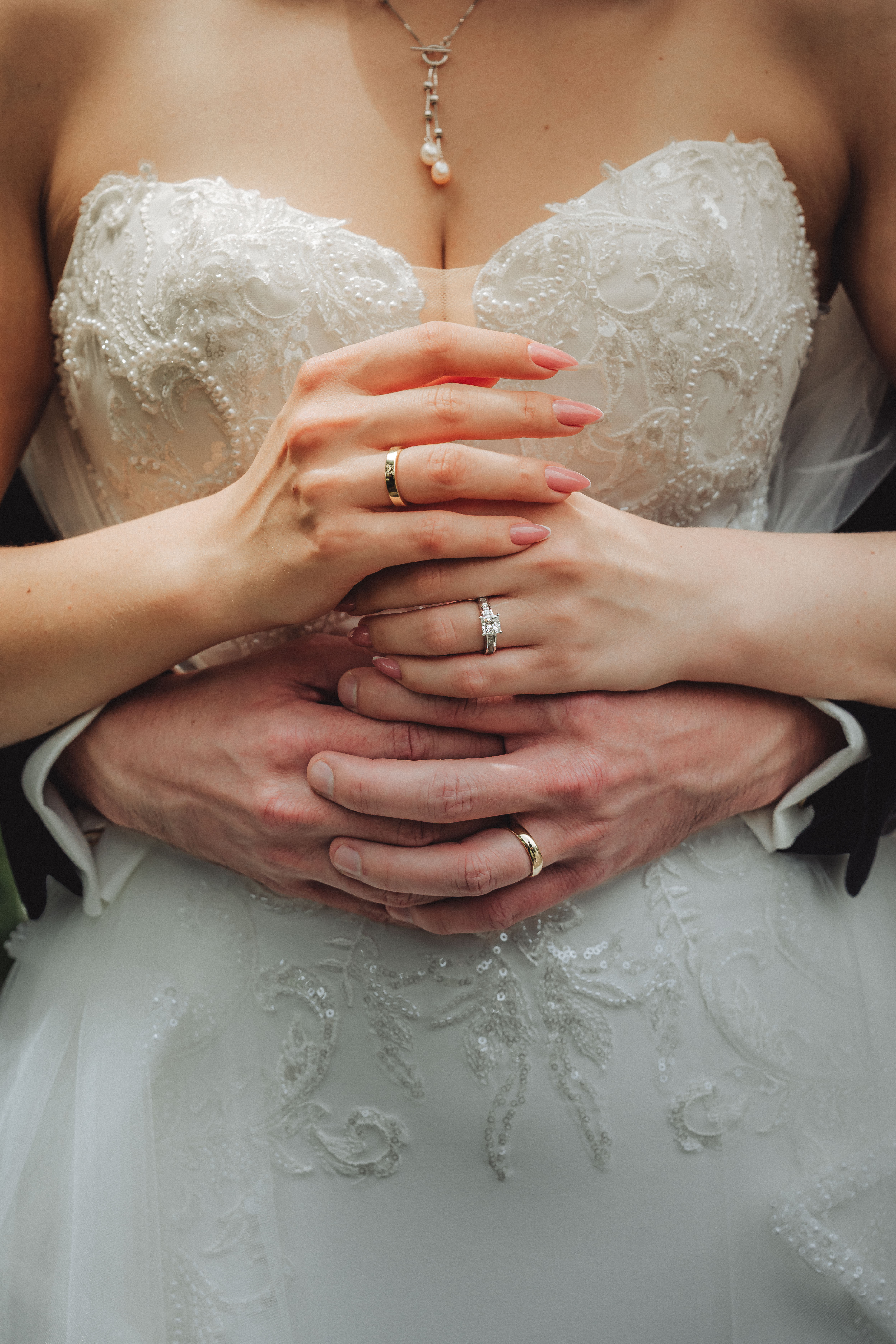 Close-up of a bride and groom’s hands with wedding rings, the bride wearing a detailed lace wedding dress.