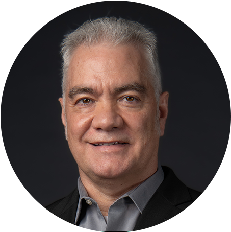 Smiling middle-aged man with gray hair wearing a dark suit and light shirt, resting his hand on a black chair against a dark background.