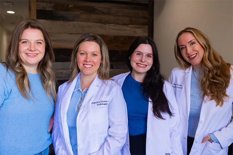 Ears4U staff standing indoors, three wearing white lab coats and one in a blue sweater, posing in front of a wooden wall.
