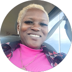 Smiling woman with short blonde hair wearing a pink top and patterned jacket inside a car.