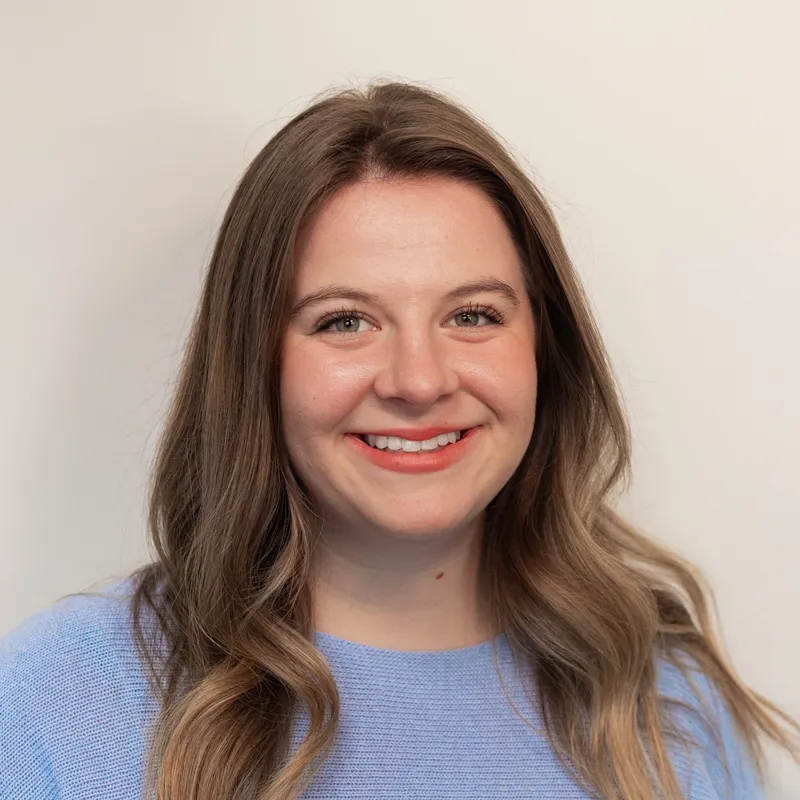 Woman with long black hair wearing glasses and a black outfit standing indoors near a white door, smiling.