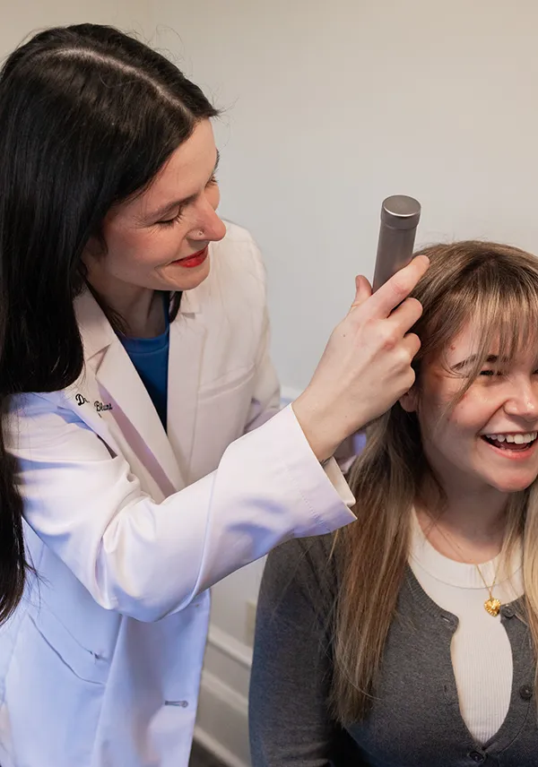 Healthcare professional smiling while examining a smiling woman's scalp with a handheld device.