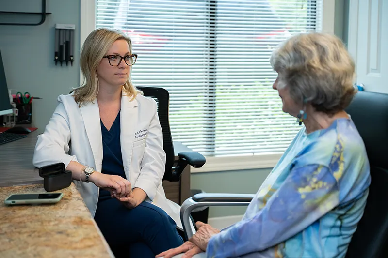 Audiologist in white coat talking with older woman seated across from her in an office with window blinds.