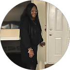 Woman with long black hair wearing glasses and a black outfit standing indoors near a white door, smiling.