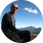 Man wearing sunglasses sitting on a rock with mountains and blue sky in the background.