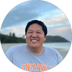 Smiling man wearing a light gray sweatshirt standing outdoors with a blurred landscape and cloudy sky in the background.