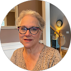 Smiling older woman with glasses and gray hair pulled back, wearing a beige patterned top, sitting indoors by a window and lamp.