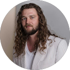 Man with long curly hair and beard wearing a white pinstripe blazer and white shirt, posing against a gray background.