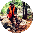 Hunter wearing an orange vest and hat posing with a deer in a forested area.