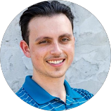 Smiling man with dark hair wearing a blue striped polo shirt in front of a gray textured wall.