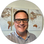 Smiling man with glasses wearing a light blue shirt, standing in front of a wall with three world map panels.