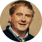 Portrait of a smiling middle-aged man with light brown hair wearing a dark sweater over a collared shirt.