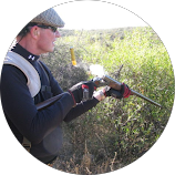 Man in outdoor gear aiming a rifle in a grassy field during daylight.