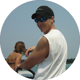 Man wearing a black cap, sunglasses, and white sleeveless shirt outdoors with a lake in the background.