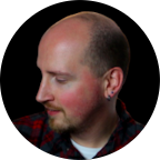 Side profile of a bald man with a trimmed beard wearing a plaid shirt and an earring.