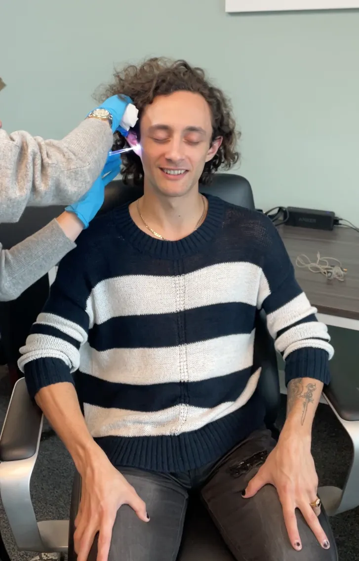 Young man sitting in a chair with eyes closed while an Audiologist wearing blue gloves cleans his ear with a lighted tool.