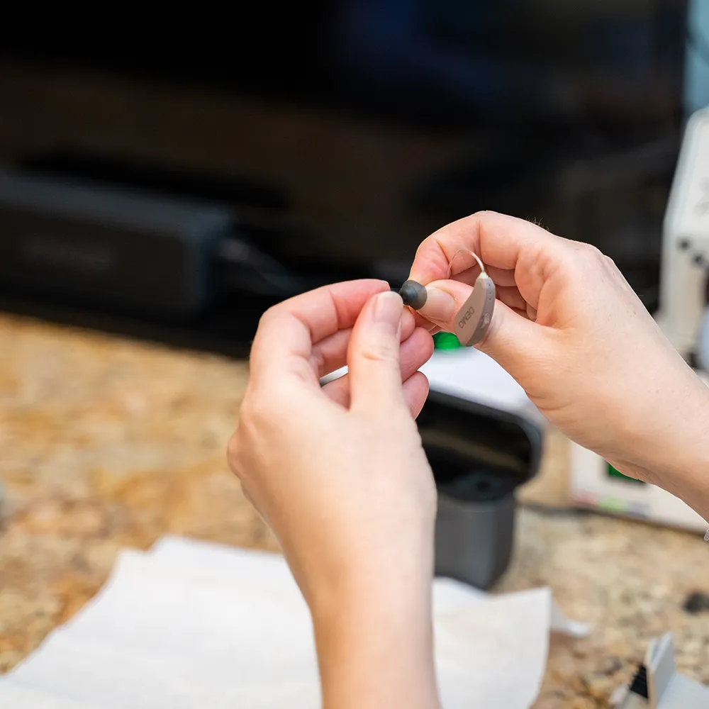 Person holding and inspecting a hearing aid over a countertop with cleaning supplies nearby.