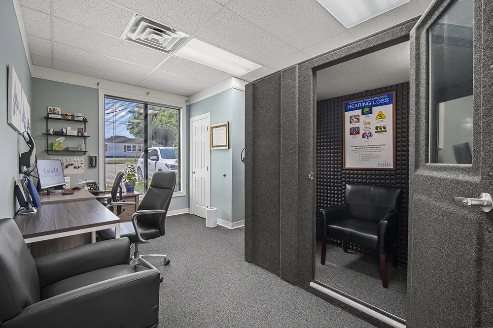 Office room with a large window, black chairs, a desk with computer and shelves, and a soundproof booth displaying a hearing loss information poster.