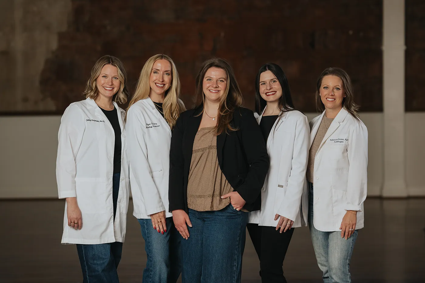 Ears 4 U team standing close together indoors, all wearing blue tops with varied hairstyles, two with long blonde hair and one with long dark hair showing tattoos on her arm.