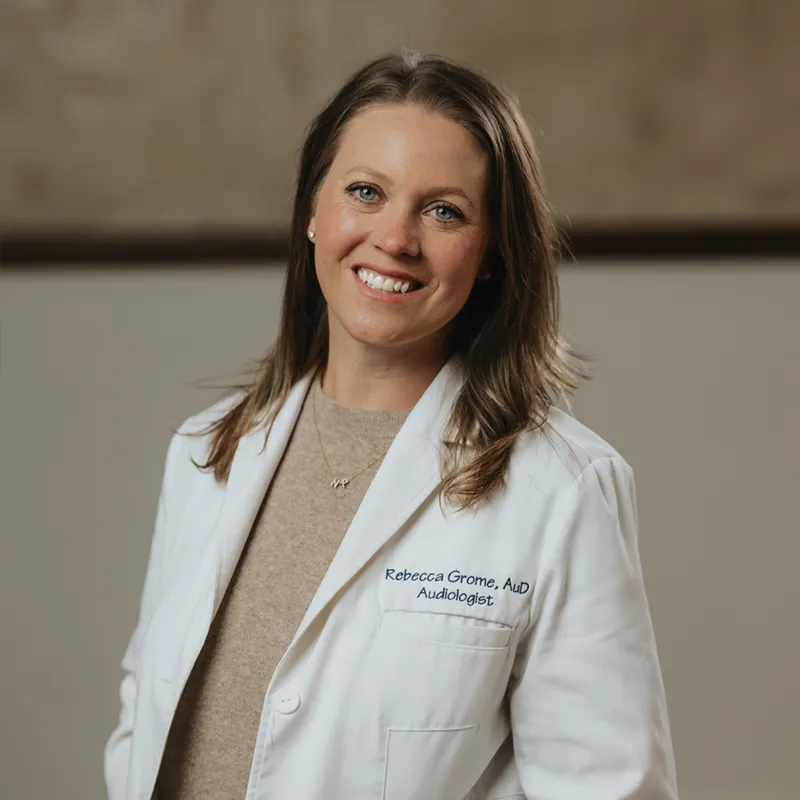 Smiling man wearing a light gray sweatshirt standing outdoors with a blurred landscape and cloudy sky in the background.