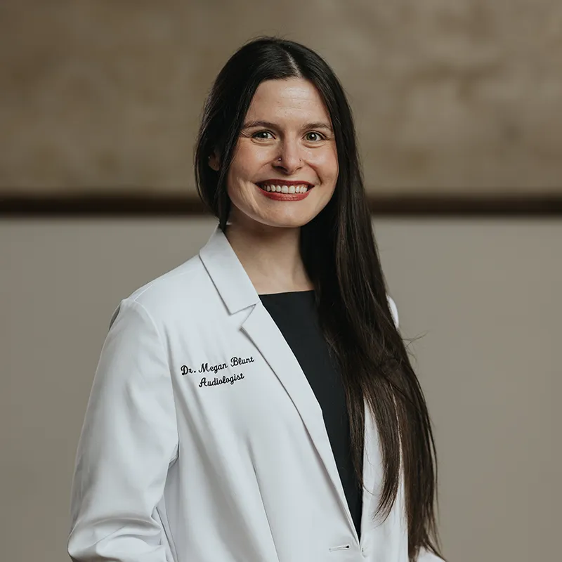 Smiling man wearing a light gray sweatshirt standing outdoors with a blurred landscape and cloudy sky in the background.