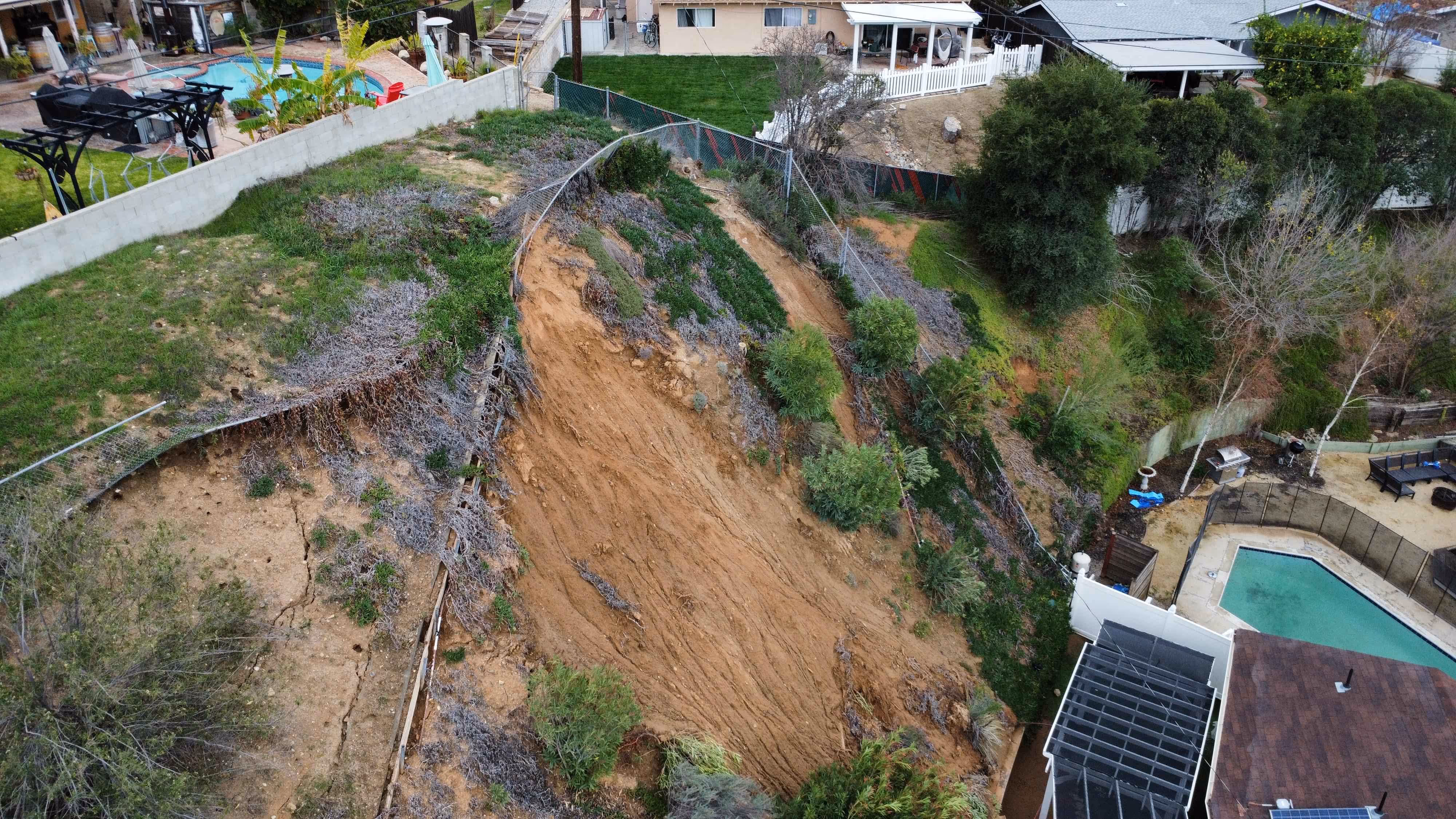 Aerial view of a residential area showing a landslide in Santa Clarita that has caused a section of a hillside with vegetation to collapse near houses and swimming pools.