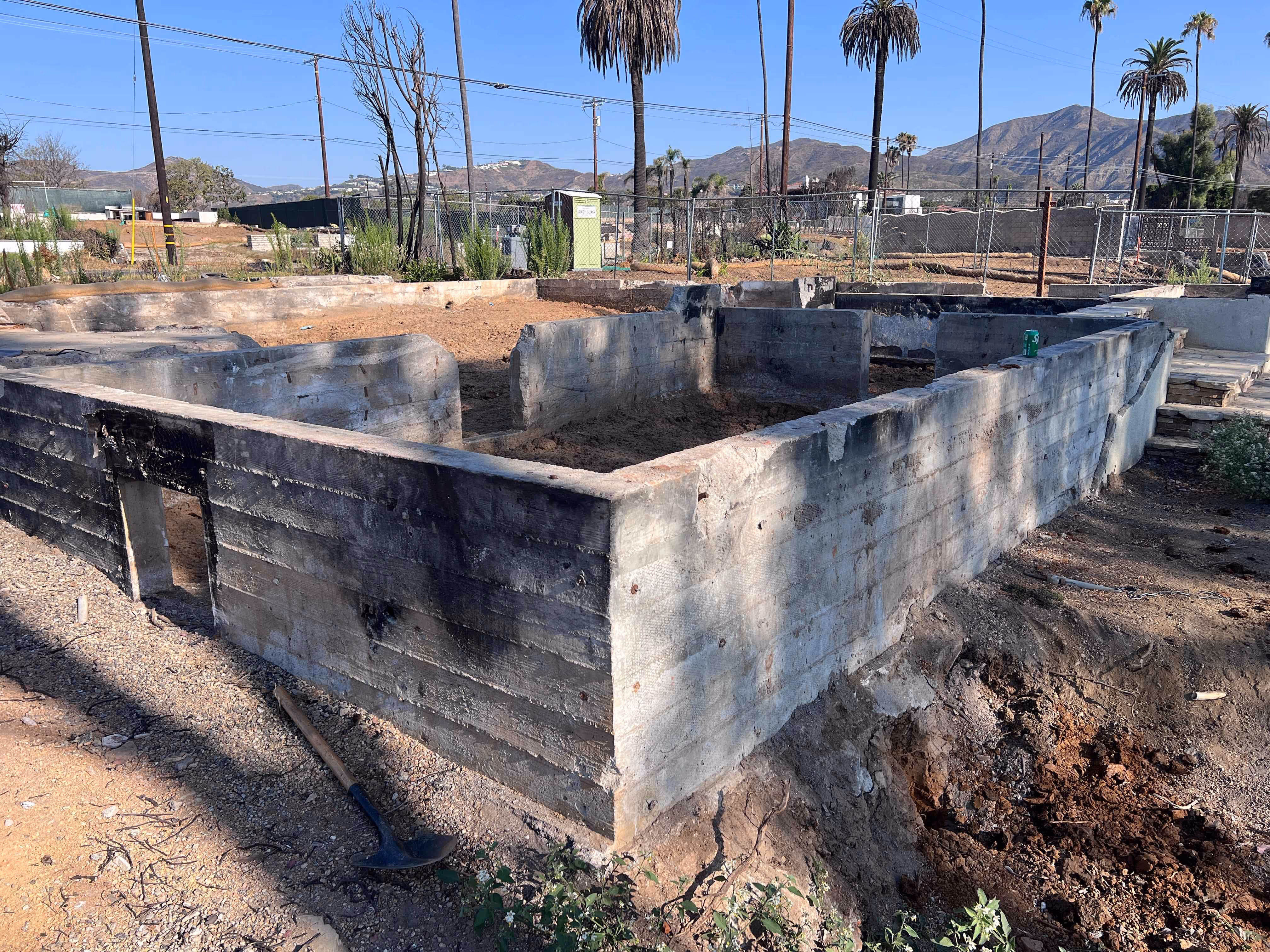 Concrete foundation walls of a building under construction in Santa Clarita with a shovel resting on the ground nearby and palm trees in the background.