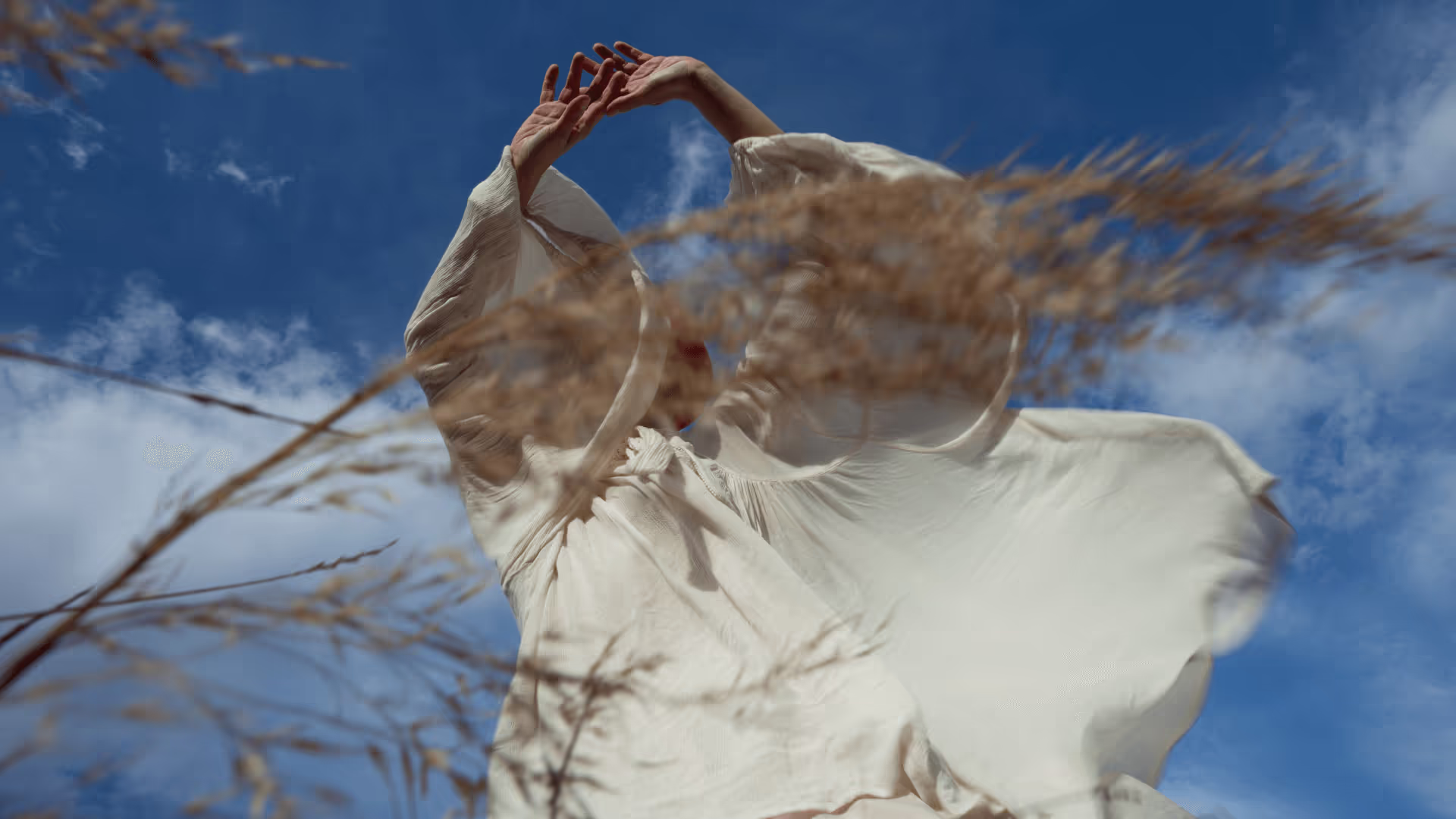 Person in a flowing white dress with arms raised, seen from below against a blue sky with clouds and blurred dry grass in the foreground.