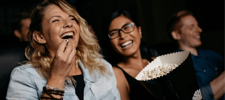 Three friends laughing and sharing popcorn while watching a movie in a theater.