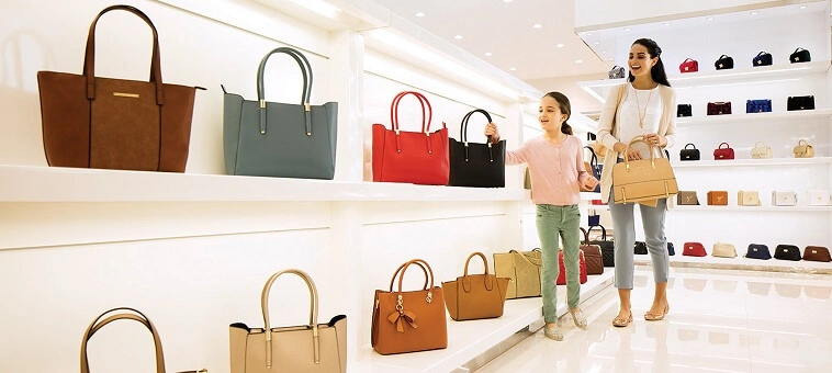 Smiling woman and girl shopping for handbags in a bright store with shelves displaying various handbags.