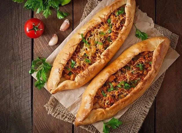 Two baked Turkish pide breads topped with minced meat, cheese, and parsley on parchment paper with tomato, garlic cloves, and parsley on wooden surface.