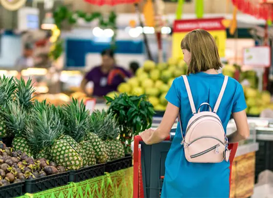 Woman in blue dress with pink backpack shopping for pineapples in a grocery store.