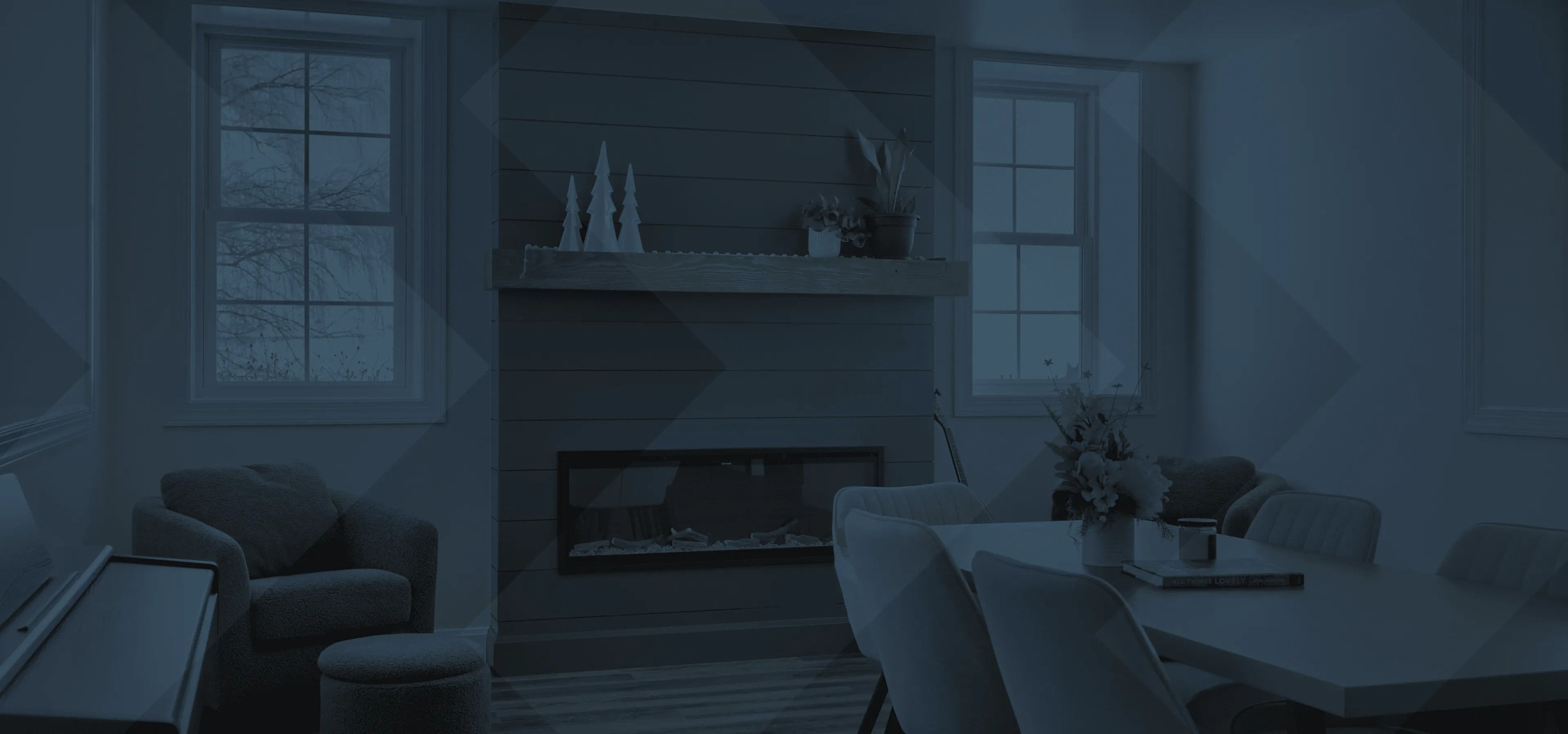 Cozy remodelled basement space with two armchairs, a modern fireplace, a dining table with chairs, flower vase, and large windows showing tree branches outside.