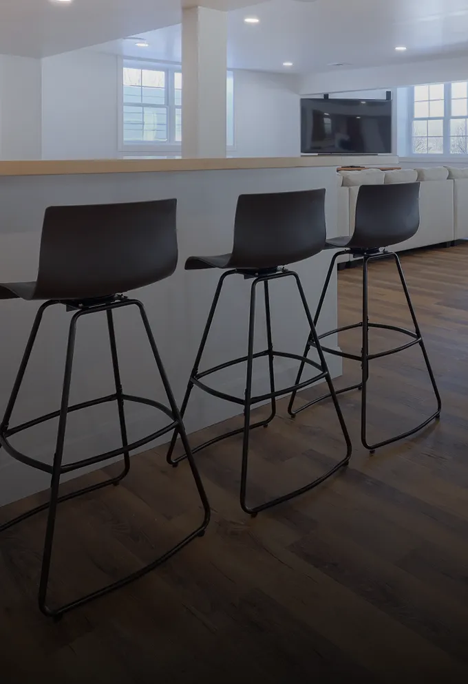 Modern kitchen bar with three black high stools on dark wood flooring and a white couch with a wall-mounted TV in the background.
