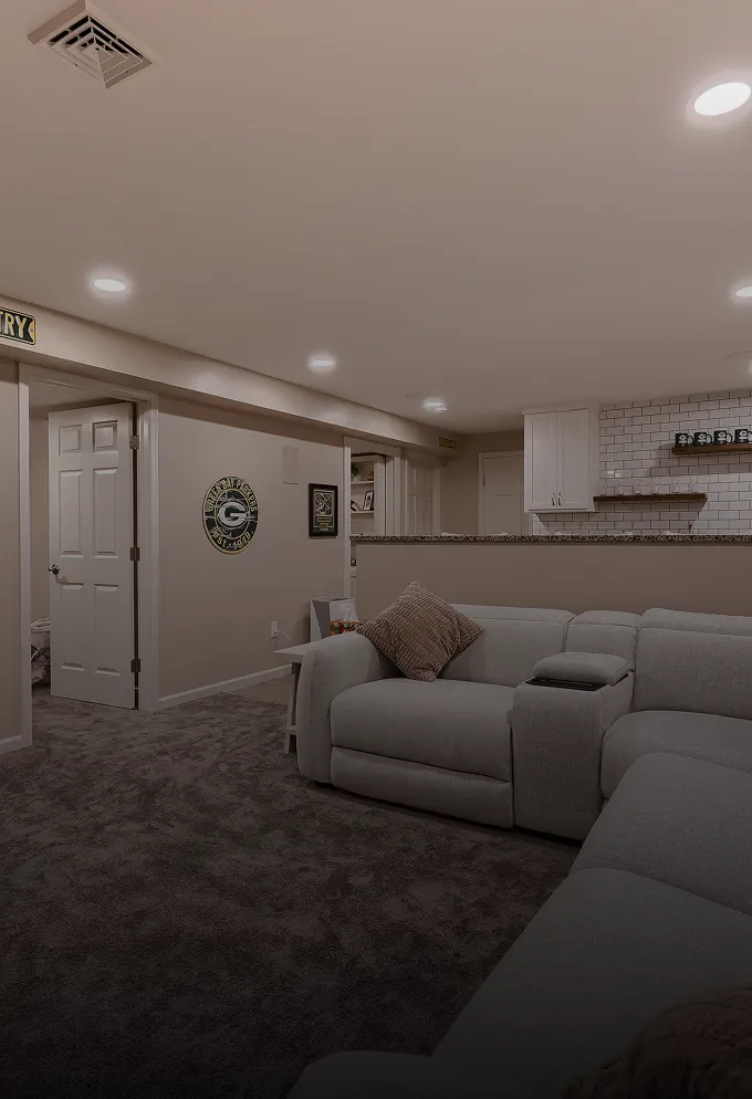 Basement living area with gray sectional sofa featuring a brown pillow, carpeted floor, and partial view of a kitchen with white subway tile backsplash.