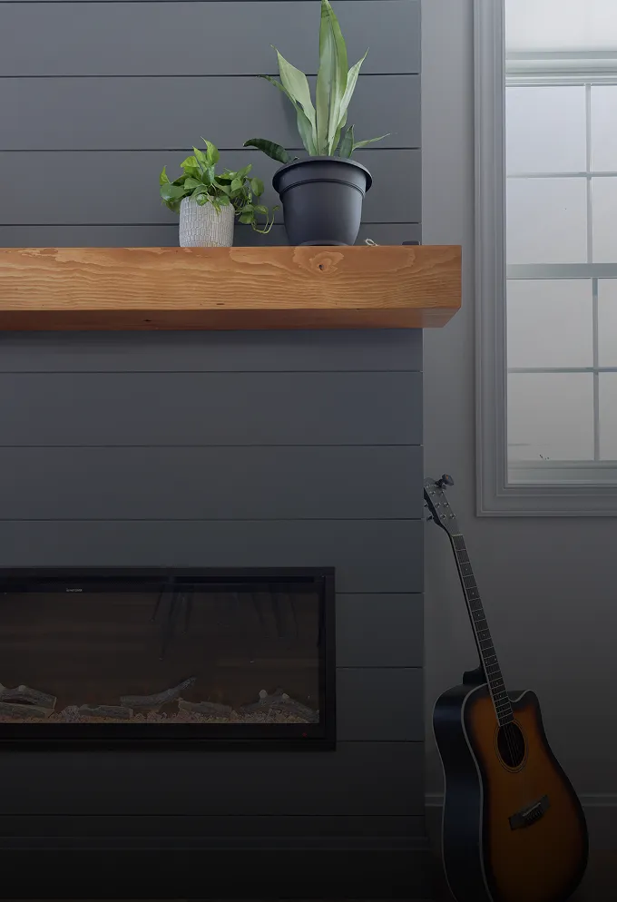 Dark grey paneled wall with a wooden mantel holding two potted plants above a modern fireplace, and an acoustic guitar leaning nearby.
