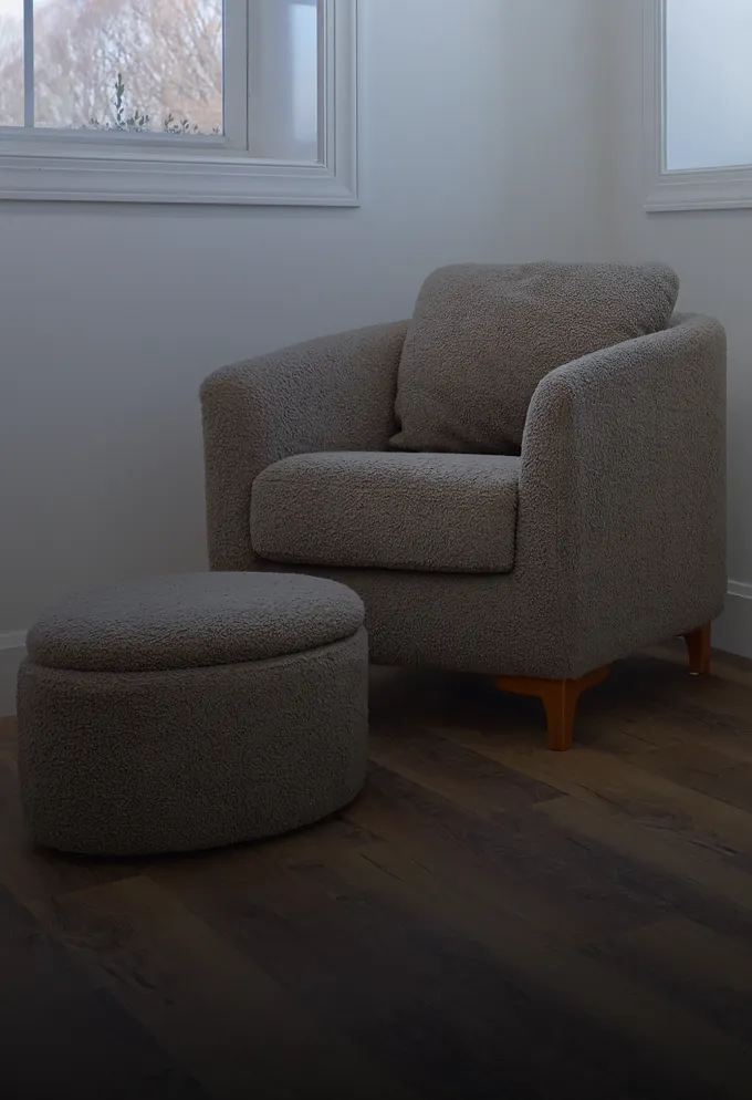 Gray upholstered armchair with matching round ottoman on wooden floor near two windows.