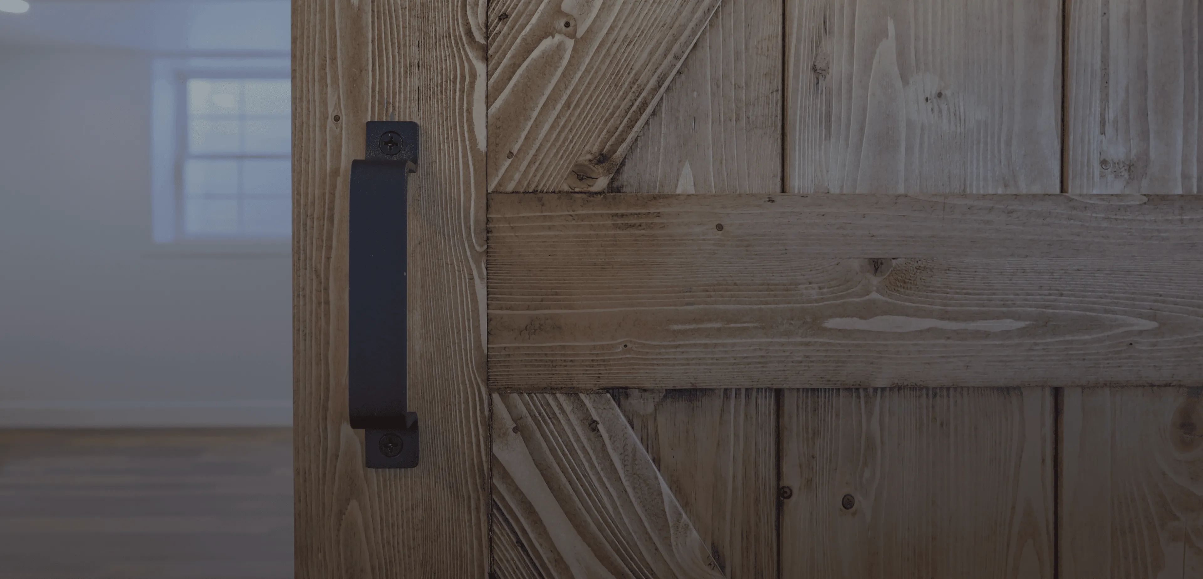 Close-up of a rustic wooden door with a black metal handle and blurred window in the background.