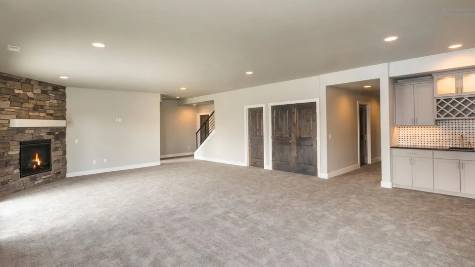 Spacious basement recreation room with light gray carpet, stone fireplace, white walls, dark wood doors, and built-in gray cabinets with a wine rack.
