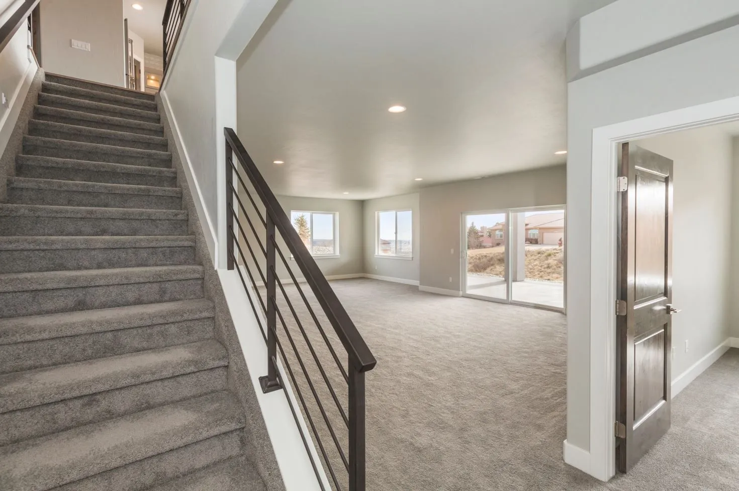 Bright, spacious carpeted basement room with large windows, sliding glass door, and carpeted stairs with dark metal railing.