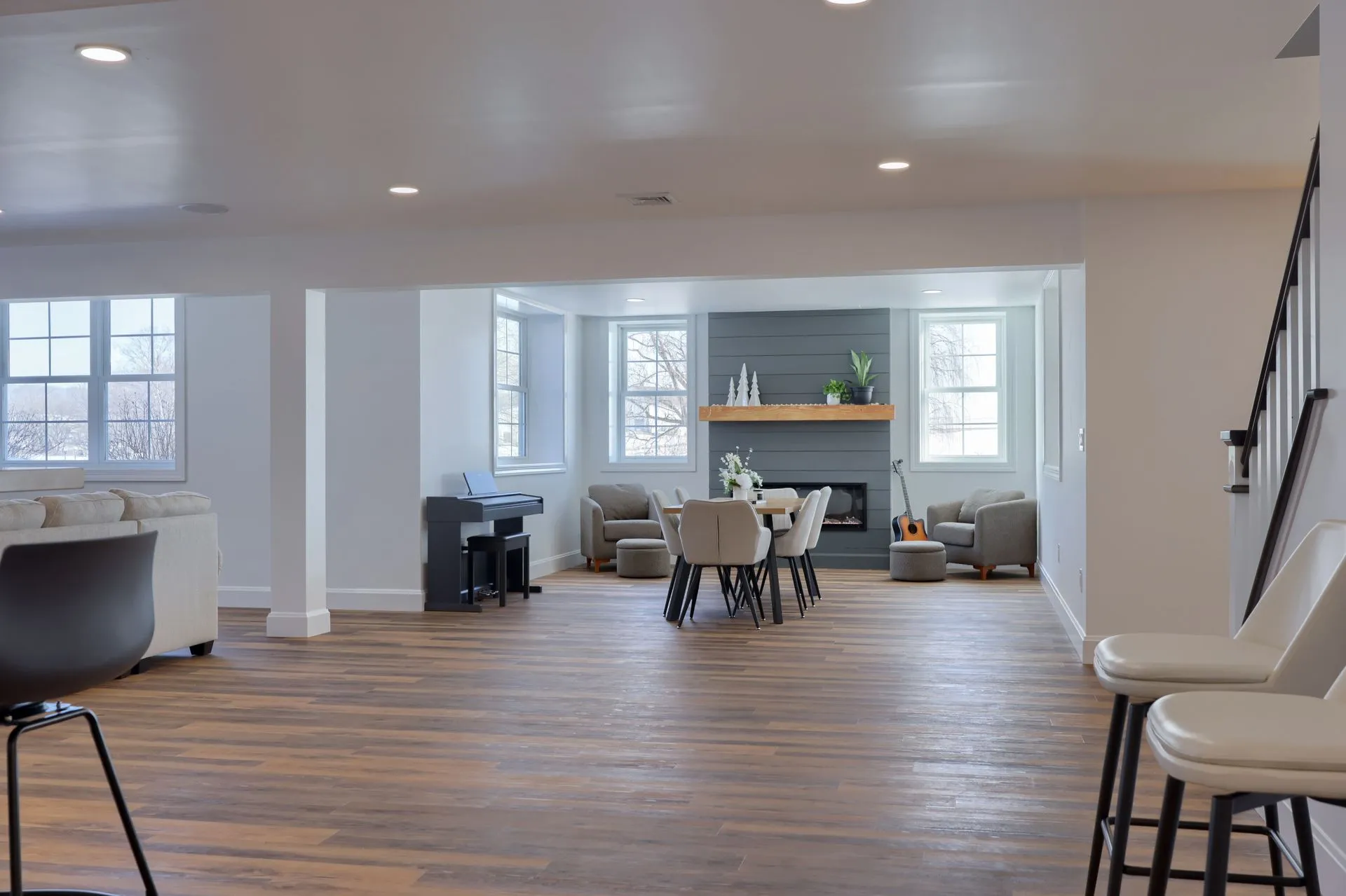 Spacious modern looking remodelled basement with wooden floor, a dining table, armchairs, a fireplace, piano, and large windows.