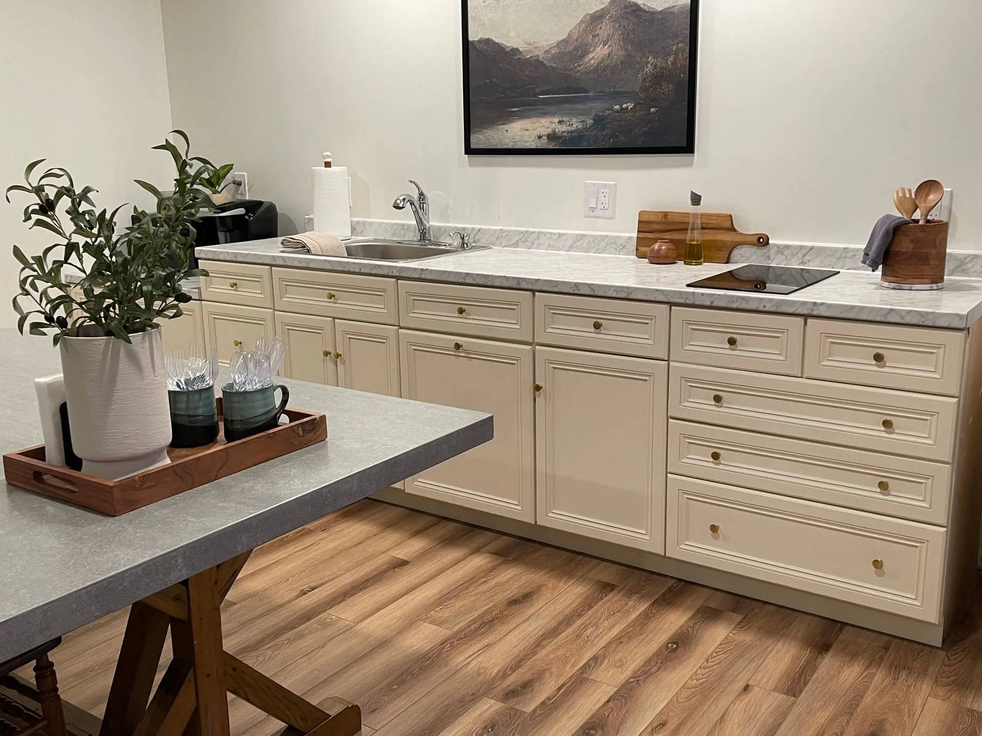Modern kitchen area with cream cabinets, marble countertop, sink, stovetop, wooden utensils, and a wooden floor, featuring a framed mountain landscape painting.