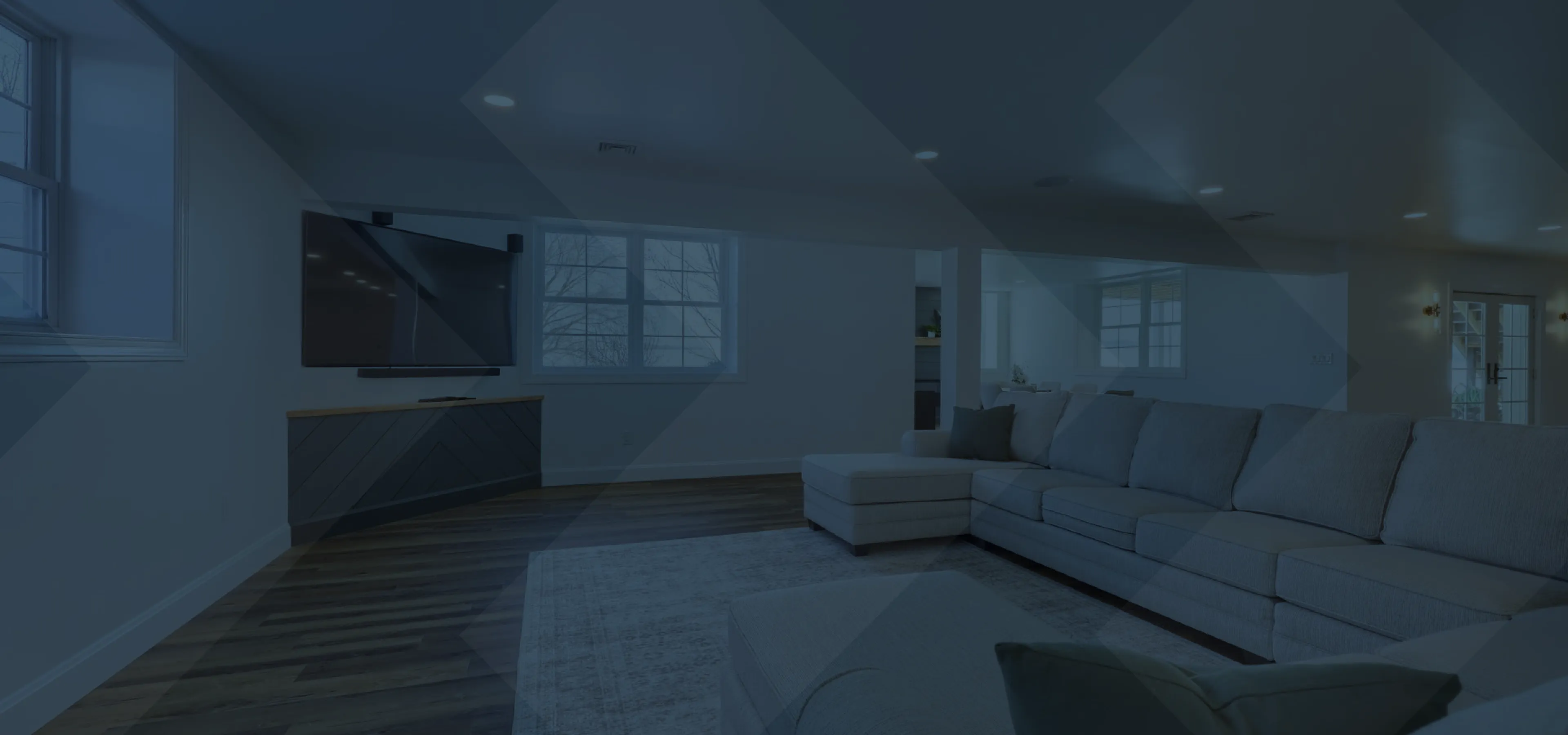 Modern remodelled basement with a large gray sectional sofa, a wall-mounted flat-screen TV, hardwood floors, and white walls with windows.