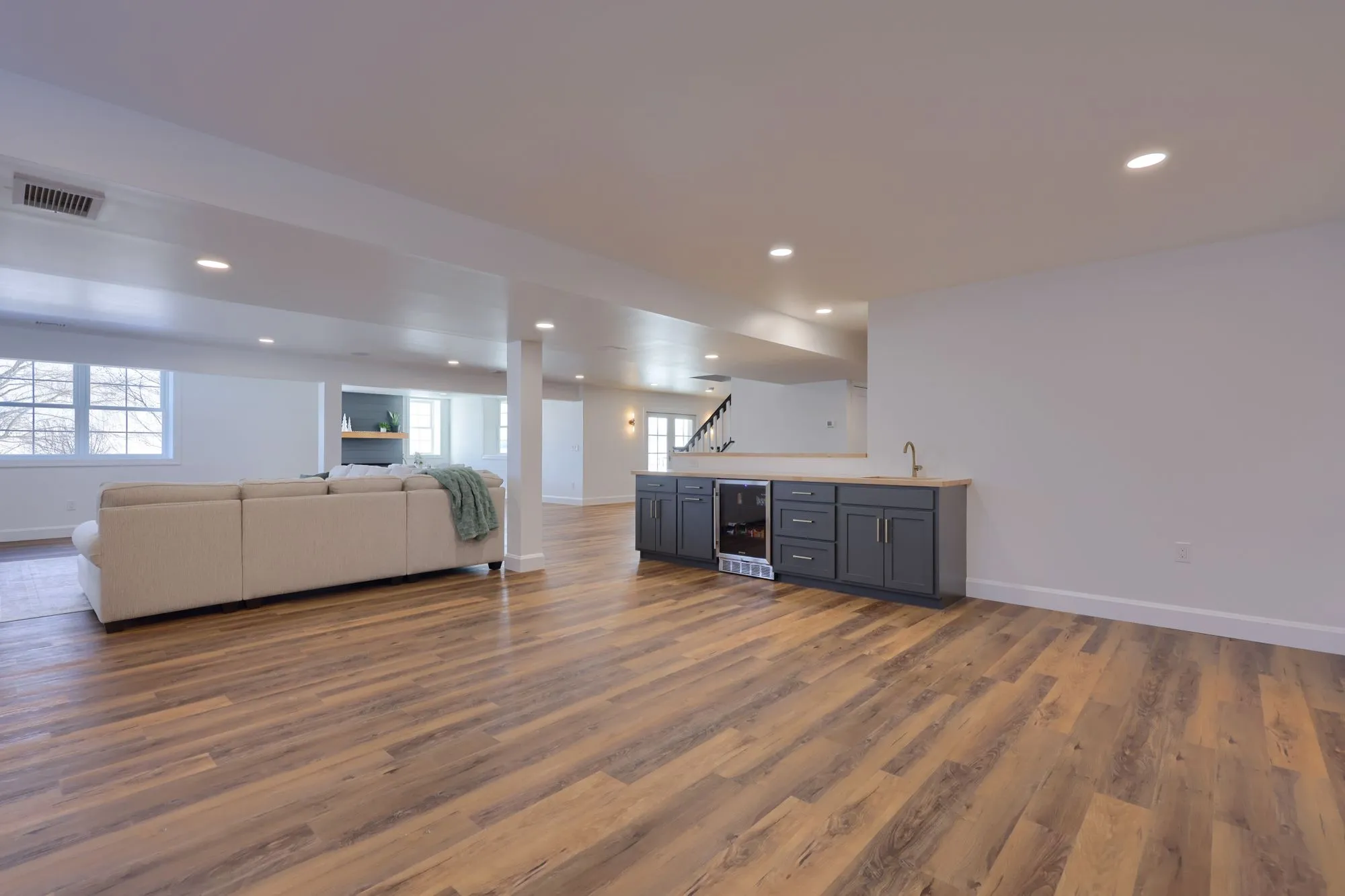 Spacious modern basement with wooden flooring, a beige sectional sofa, and a wet bar with dark cabinets and a small fridge.